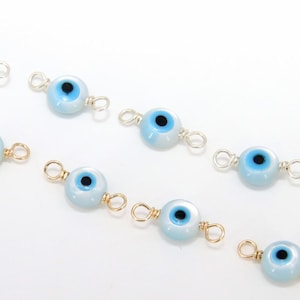 Puede incluir: Varios amuletos de ojo turco de color azul claro y dorado están dispersos sobre una superficie blanca. Cada amuleto presenta un ojo azul con una pupila negra, conectado por bucles dorados. Los amuletos están hechos a mano por A Girls Gems.