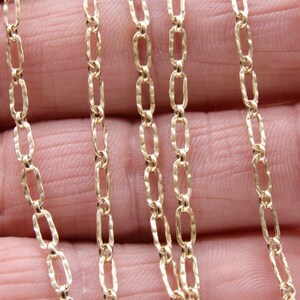 May include: A close-up of a gold chain necklace with a textured, oval-shaped link. The chain is displayed on a hand, with multiple strands of the chain visible.