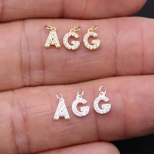 May include: Three gold and three silver letter charms spelling "AGG". Each charm is a capital letter with a loop at the top for attaching to a chain. The letters are encrusted with small, clear gemstones.
