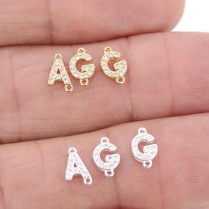 May include: Close-up of three sets of letter charms. Two sets are gold-toned, and one set is silver-toned. Each set includes the letters A, G, and G, encrusted with small, clear crystals. The charms are for jewelry making.