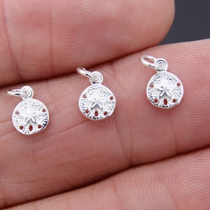 May include: Three silver sand dollar charms with a detailed design. The charms are shown on a hand, ready to be added to jewelry.