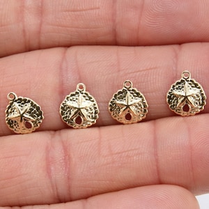 May include: Five gold-colored sand dollar charms with a star design. The charms are on a hand.