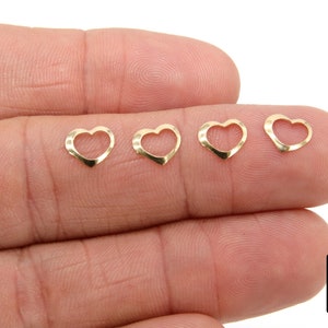 May include: Four gold-colored heart-shaped charms with an open center. The charms are displayed on a finger.