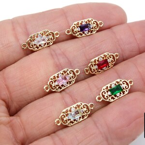 May include: Six gold-tone connector charms with ornate filigree designs and a variety of coloured gemstones in the centre. The gemstones are purple, red, pink, green, and light blue.