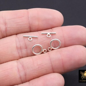 May include: Close-up of silver-colored jewelry clasps. The clasps include two circular rings with attached loops and two T-shaped bars with loops. The clasps are small and delicate.