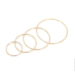 May include: Four gold-colored hoop earrings of varying sizes are arranged diagonally on a white background. The hoops are thin and circular, with a shiny finish. The smallest hoop is on the left, and the largest is on the right. The logo of "A Girls Gems" is in the bottom right corner.