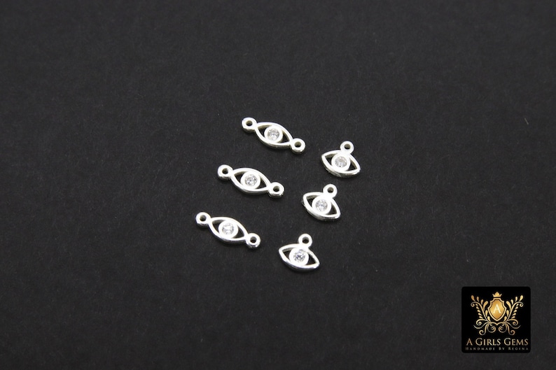 May include: Six silver eye charm pendants with clear rhinestone accents. The charms are oval shaped with a small loop at the top for attaching to jewelry.