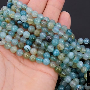 May include: Strands of round, polished gemstone beads in shades of blue, green, and cream. The beads are held in a hand, showcasing their varying colors and textures. The beads are likely for jewelry making.