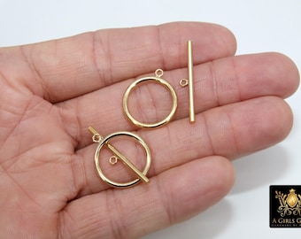 May include: Two gold-toned metal toggle clasps, one with a round ring and the other with a straight bar. The clasps are shown on a hand.