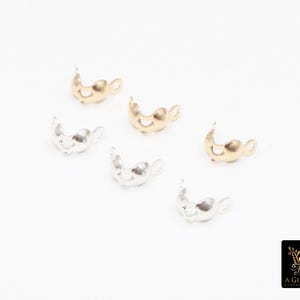 May include: Six small, decorative jewelry findings in gold and silver tones are scattered on a white surface. The findings have a curved, open design with a small loop on one end. The logo of "A Girls Gems" is in the bottom right corner.
