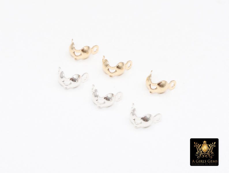 May include: Six small, decorative jewelry findings in gold and silver tones are scattered on a white surface. The findings have a curved, open design with a small loop on one end. The logo of "A Girls Gems" is in the bottom right corner.