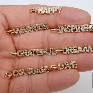 May include: A set of gold charm necklaces with various words set in diamonds, including "Happy", "Warrior", "Inspire", "Grateful", "Dream", "Courage", and "Love".