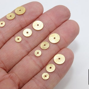 May include: Assortment of gold-colored metal round sequins of varying sizes. The sequins have a textured surface and a hole in the center.