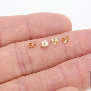 May include: Four gold earring backs on a hand. The backs are small and round with a hole in the center.