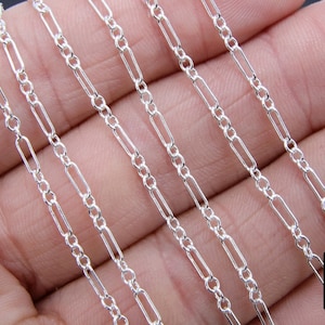 May include: Five strands of silver chain, each with a different length, are laid out on a hand. The chain is a delicate, fine, and flat oval link chain.