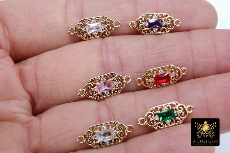 May include: Five gold-tone metal connector charms with different coloured gemstones. Each charm features a decorative filigree design and a rectangular gemstone in the centre. The gemstones are clear, purple, red, pink, and green.