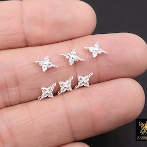 May include: Six small, silver-toned, four-petal flower-shaped stud earrings with clear, sparkling gemstones. The earrings are displayed on a light-coloured background. The earrings are delicate and elegant.