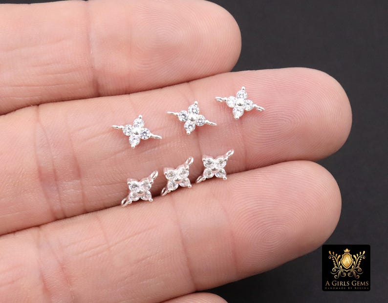 May include: Six small, silver-toned, four-petal flower-shaped stud earrings with clear, sparkling gemstones. The earrings are displayed on a light-coloured background. The earrings are delicate and elegant.