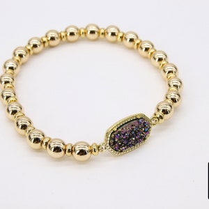 May include: Gold beaded bracelet with a purple and iridescent gemstone charm. The charm is oval shaped and has a gold setting.