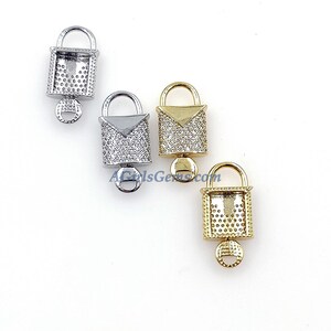 May include: Three lock charms with a square shape and a small keyhole. The charms are made of metal and have a clear crystal-like finish. The charms are in silver, gold, and a mixed silver and gold color.