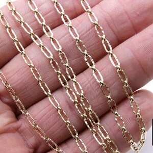May include: Close-up of a gold chain necklace with a textured, oval-shaped link. The chain is shown on a hand, with multiple strands layered on top of each other.