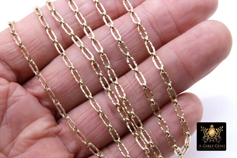 May include: Close-up of a gold chain necklace with a textured, oval-shaped link. The chain is shown on a hand, with multiple strands layered on top of each other.