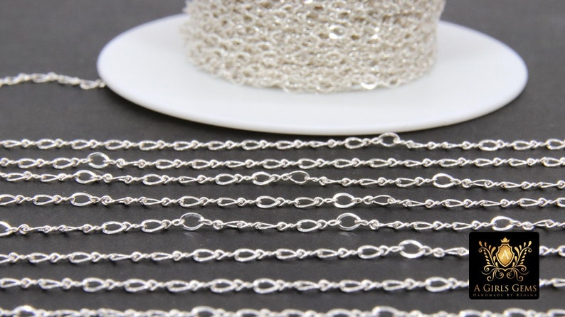 May include: A silver chain with a twisted oval link design. The chain is coiled on a white spool and laid out in rows on a black surface.