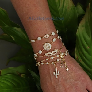 May include: A set of five gold bracelets with white enamel charms. The charms include a circle with a diamond-like design, a mask, and a cactus. The bracelets are layered on a person's wrist.