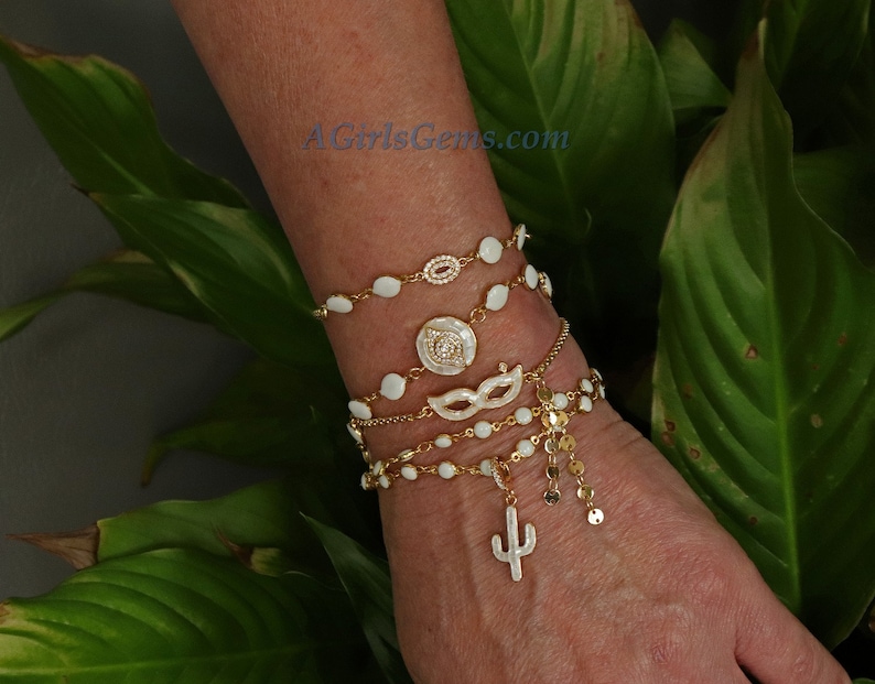 May include: A set of five gold bracelets with white enamel charms. The charms include a circle with a diamond-like design, a mask, and a cactus. The bracelets are layered on a person's wrist.
