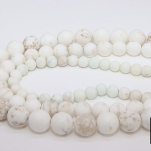 May include: A strand of white howlite beads with brown speckles. The beads are round and smooth, and they are arranged in a circle. The beads are of varying sizes.