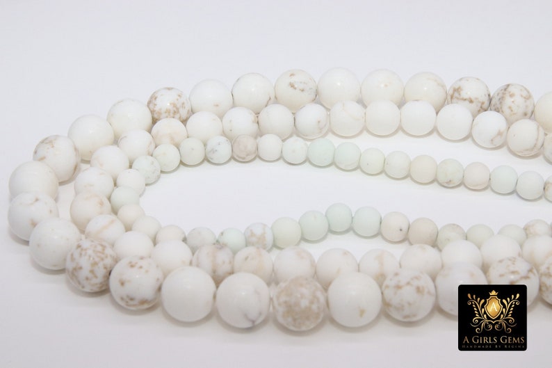 May include: A strand of white howlite beads with brown speckles. The beads are round and smooth, and they are arranged in a circle. The beads are of varying sizes.