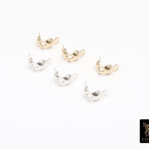 May include: Six metal jewelry findings in gold and silver tones are scattered on a white surface. The findings are shaped like a curved cup with a small loop on each side. The logo for A Girls Gems is in the bottom right corner.