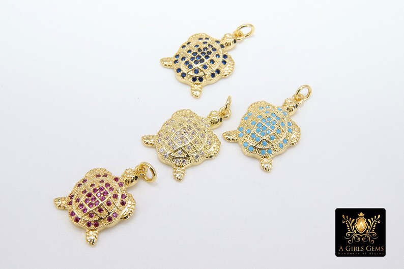 Op de afbeelding: Vier gouden schildpadbedels met verschillende gekleurde kristallen. De schildpadden zijn allemaal even groot en even gevormd, maar hebben verschillende gekleurde kristallen op hun schilden. De kristallen zijn blauw, wit, turquoise en roze.