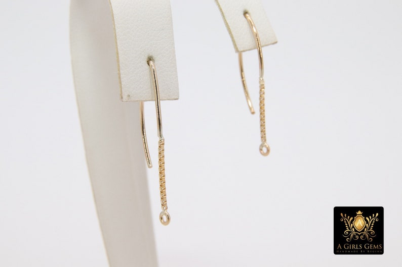 Pu&ograve; includere: Un paio di orecchini pendenti in oro con un delicato design a catena. Gli orecchini sono esposti su un espositore per gioielli bianco.