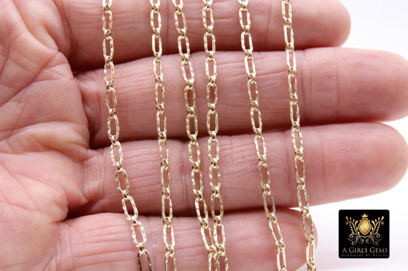 May include: A close-up of a gold chain necklace with a textured, oval-shaped link. The chain is displayed on a hand, with multiple strands of the chain visible.