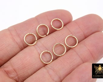 May include: A close-up of six gold-colored jump rings with a textured surface. The rings are arranged in a semi-circle on a hand.