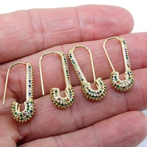 May include: Four gold safety pin earrings with colorful cubic zirconia stones. The earrings are on a hand.
