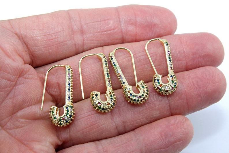 May include: Four gold safety pin earrings with colorful cubic zirconia stones. The earrings are on a hand.