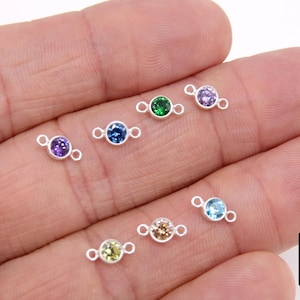 May include: A collection of small, silver-colored gemstone connectors with various colored gemstones, including purple, green, blue, and yellow. Each connector has a small loop on either side of the gemstone. The gemstones are set in a bezel setting.
