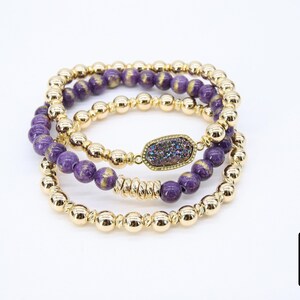 May include: Three gold bead bracelets with purple and gold beads. The middle bracelet has a large, flat, iridescent purple stone charm.