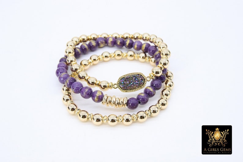 May include: Three gold bead bracelets with purple and gold beads. The middle bracelet has a large, flat, iridescent purple stone charm.
