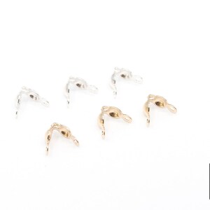 May include: Six metal jewelry findings in gold and silver tones are scattered on a white surface. The findings have a curved shape with a loop at one end and a pointed tip at the other. The logo of "A Girls Gems" is in the bottom right corner.