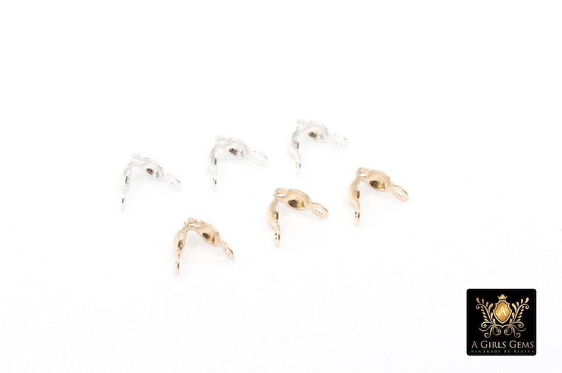 May include: Six metal jewelry findings in gold and silver tones are scattered on a white surface. The findings have a curved shape with a loop at one end and a pointed tip at the other. The logo of "A Girls Gems" is in the bottom right corner.