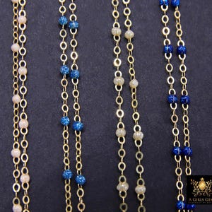 以下が含まれることがあります： さまざまな色のビーズが付いた、金色のチェーンネックレスのクローズアップ。ライトピンク、ブルー、オフホワイトなど。ネックレスは暗い背景に表示されています。「A Girls Gems」のロゴが右下に表示されています。