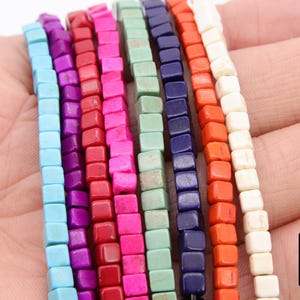 May include: Close-up of several strands of square beads in various colors, including turquoise, purple, red, pink, green, navy, orange, and white. The beads are displayed against a hand.