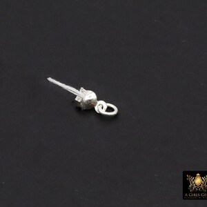 May include: A small, silver-colored jewelry finding, likely a clasp or connector, against a dark background. It features a rounded bead-like element, a small loop, and a fine, white thread extending from the top. The piece appears to be delicate and detailed.