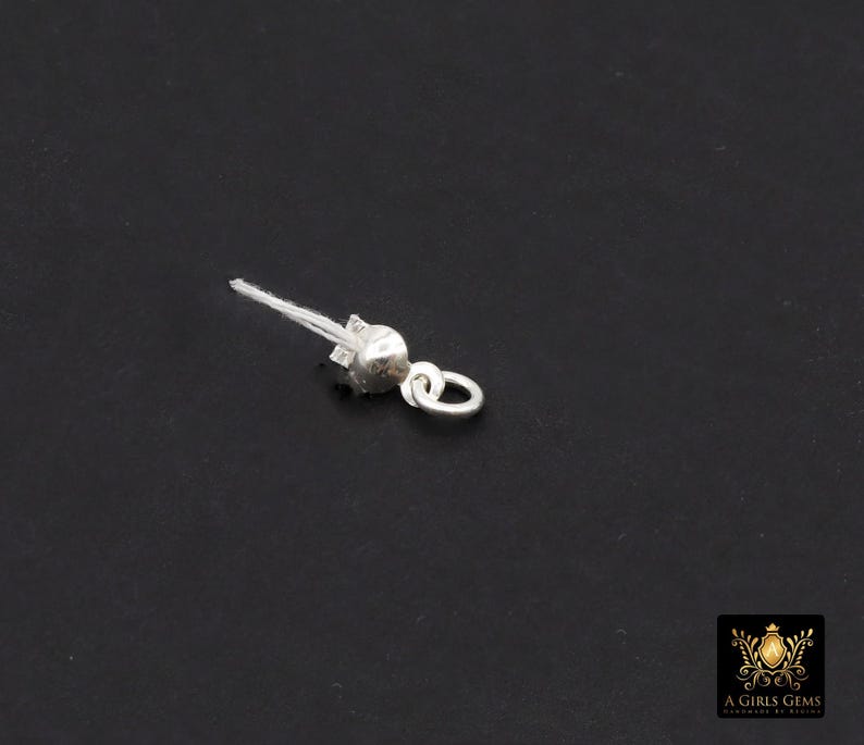 May include: A small, silver-colored jewelry finding, likely a clasp or connector, against a dark background. It features a rounded bead-like element, a small loop, and a fine, white thread extending from the top. The piece appears to be delicate and detailed.