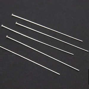 Puede incluir: Cinco alfileres de cabeza plateada, cada uno con una cabeza redondeada, están dispuestos sobre una superficie negra. Los alfileres son rectos y delgados, y todos tienen la misma longitud.