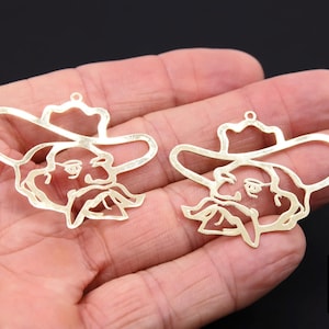 May include: Two gold-colored metal cowboy-themed earring charms. Each charm features a cutout design of a cowboy's face wearing a hat and a long mustache. The charms are held in a hand against a dark background.