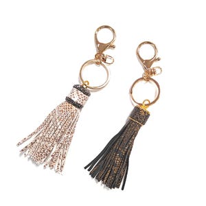 May include: Two gold keychains with tassel charms. One keychain has a white tassel with black accents and the other has a black tassel with gold accents.